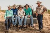 Five people dressed in rural outfits and hats sit on the back of a ute.