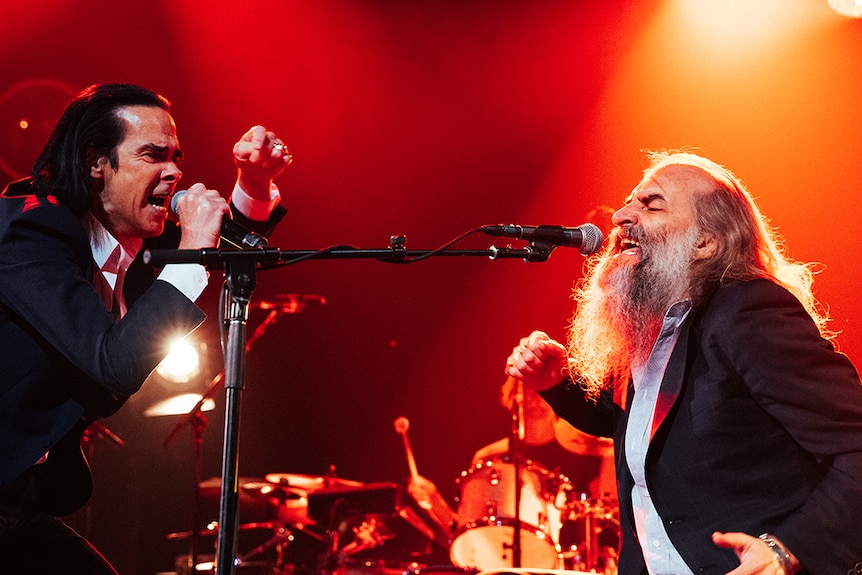 Nick Cave and Warren Ellis sing passionately into microphones on stage. Both wear suits. Ellis is bearded. Lighting is orange.