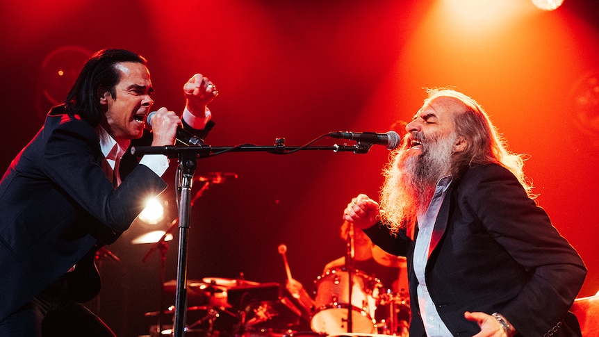 Nick Cave and Warren Ellis sing passionately into microphones on stage. Both wear suits. Ellis is bearded. Lighting is orange.