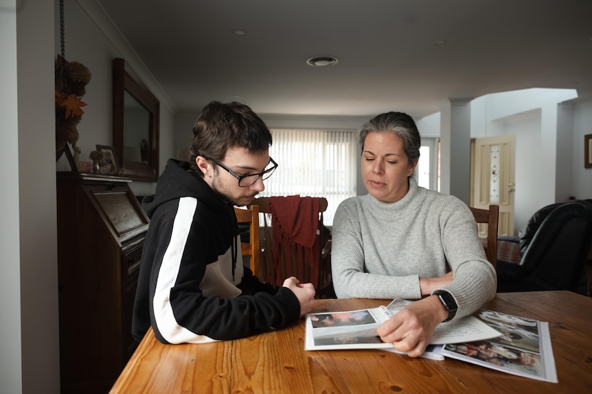 A woman and a teenager sit at a table in a family home, looking at a photo album.She is turning a page.