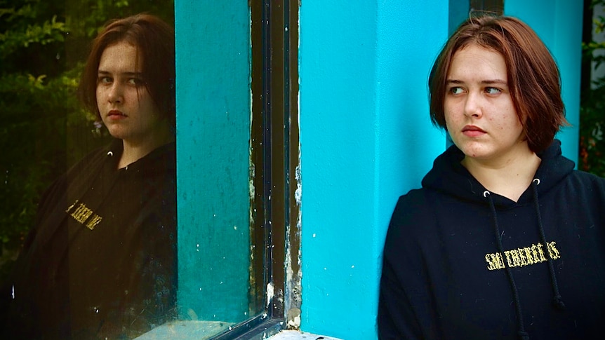 Rhiannon, wearing a black hoodie, stands against a blue wall. She is looking into a window. Her image is reflected back at her.