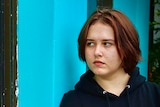 Rhiannon, wearing a black hoodie, stands against a blue wall. She is looking into a window. Her image is reflected back at her.