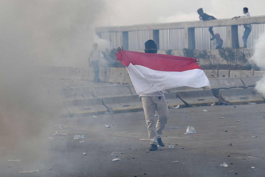 A protester holds Indonesian flag as riot police fire tear gas during a riot.