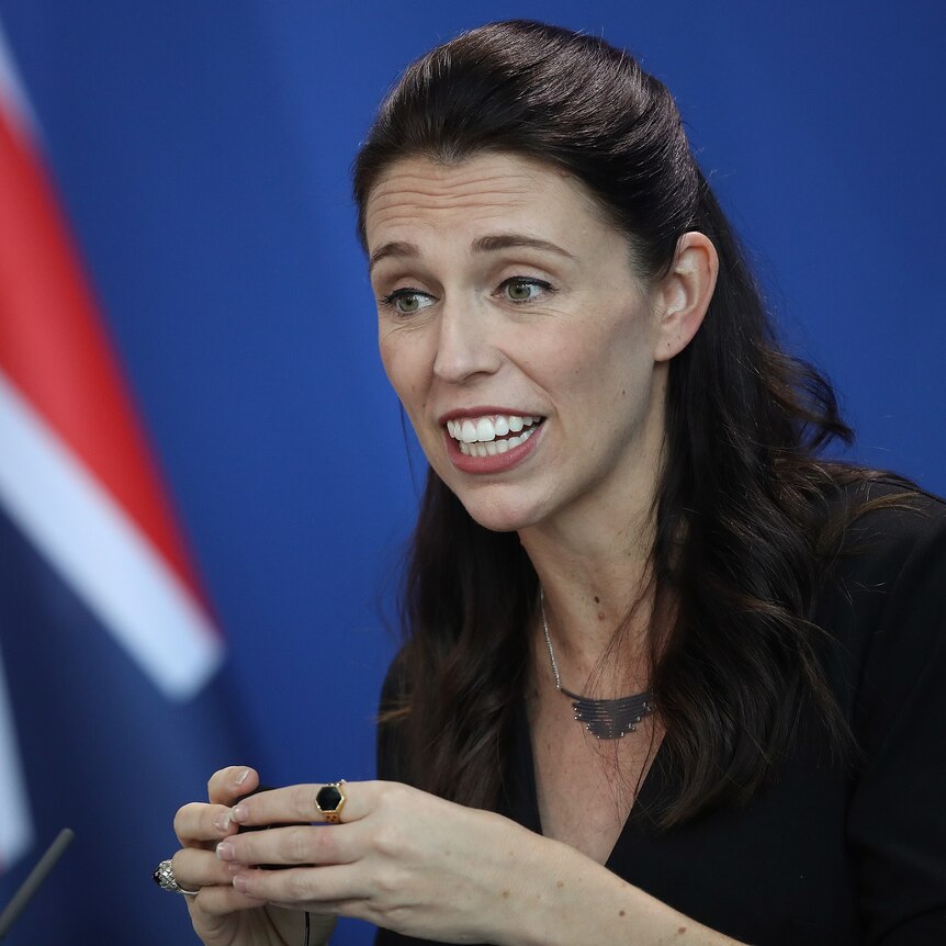 New Zealand Prime Minister Jacinda Ardern speaks to the media, with her country's flag in the background.