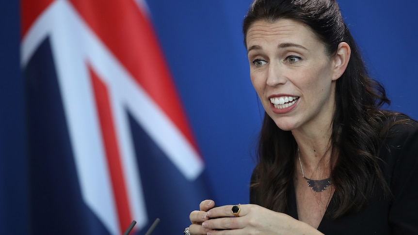 New Zealand Prime Minister Jacinda Ardern speaks to the media, with her country's flag in the background.