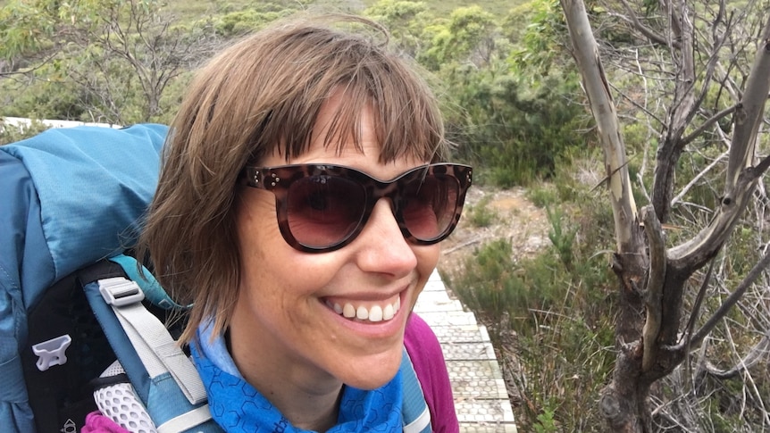 A smiling woman wearing sunglasses with a natural landscape behind her.