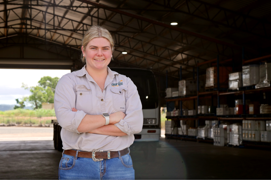 a young woman stands with her arms crossed and smiling at the camera, she's in an industrial shed stocked with farming products