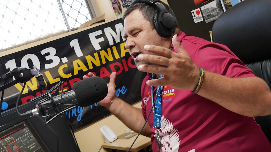 A man wearing a maroon shirt wearing headphones sitting behind a radio desk speaks into a microphone and gestures.
