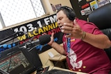 A man wearing a maroon shirt wearing headphones sitting behind a radio desk speaks into a microphone and gestures.