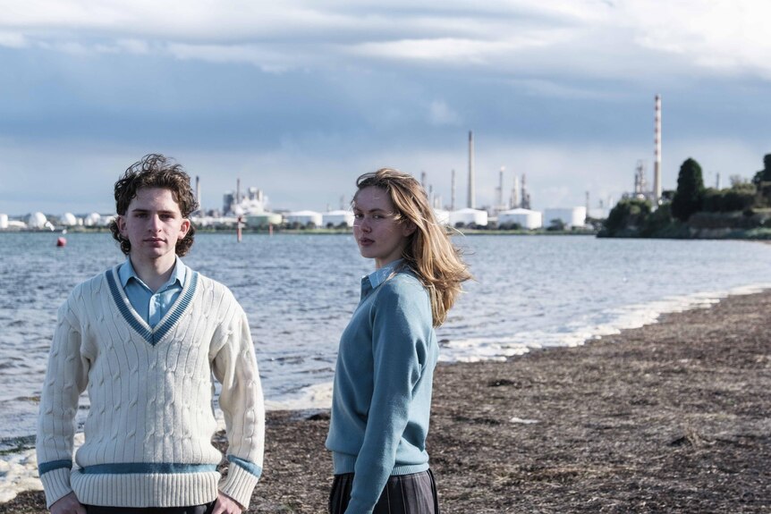 Dan and Millie stand in their school uniforms on an overcast beach, with the oil refinery visible in background.