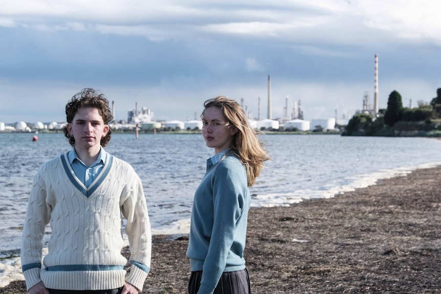 Dan and Millie stand in their school uniforms on an overcast beach, with the oil refinery visible in background.