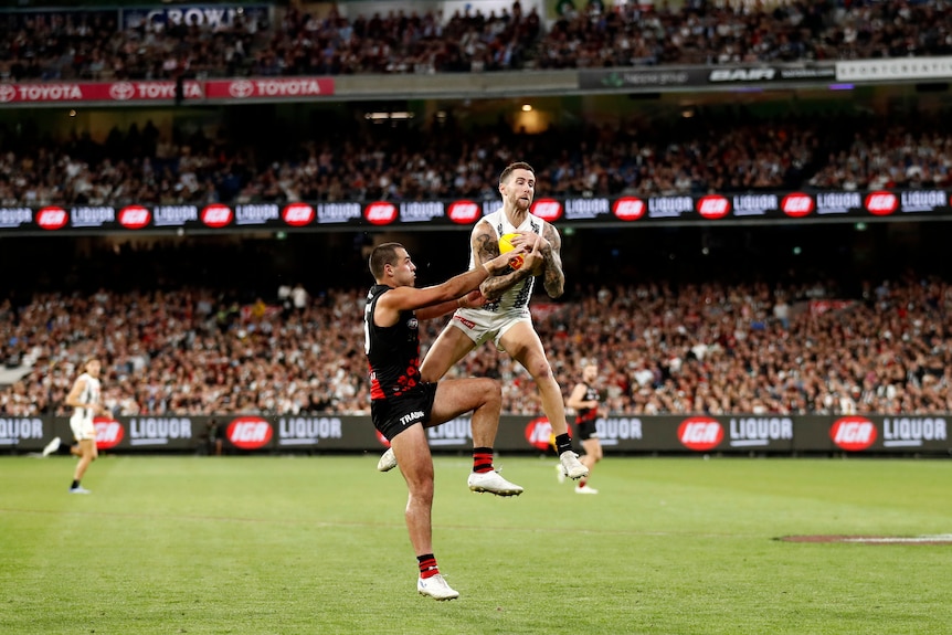 Collingwood AFL player Jeremy Howe leaps in the air and grabs the ball to his chest ahead of an Essendon opponent.