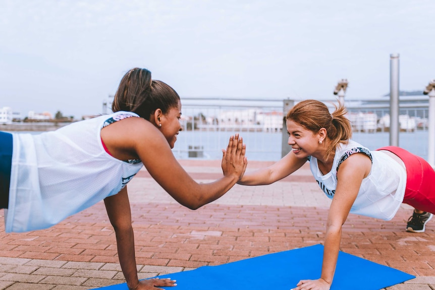 Two women in activewear touch hands while holding a plank position on yoga mats on a rooftop.