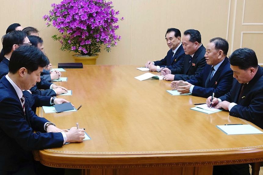 South and North Korean officials sit opposite each other at a table talking and taking notes.