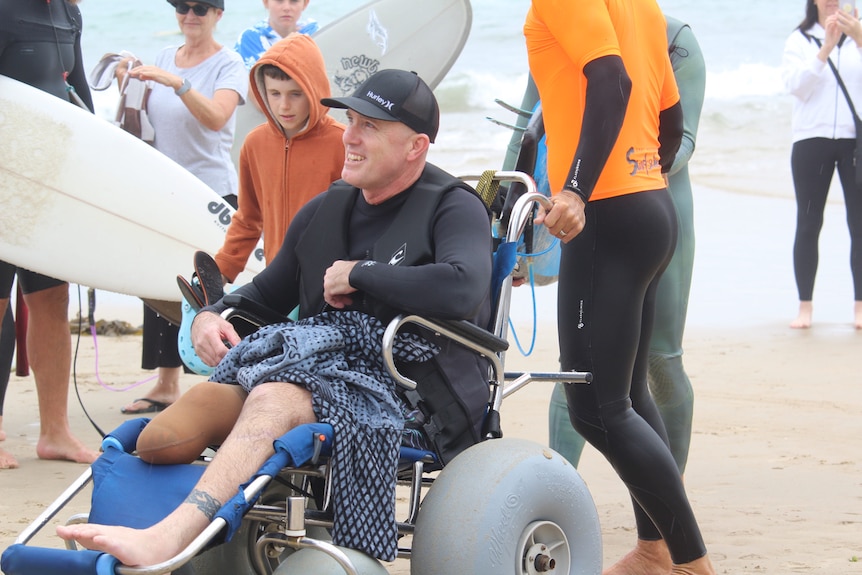 A man sitting in a beach wheelchair, wearing a wetsuit, at a beach.