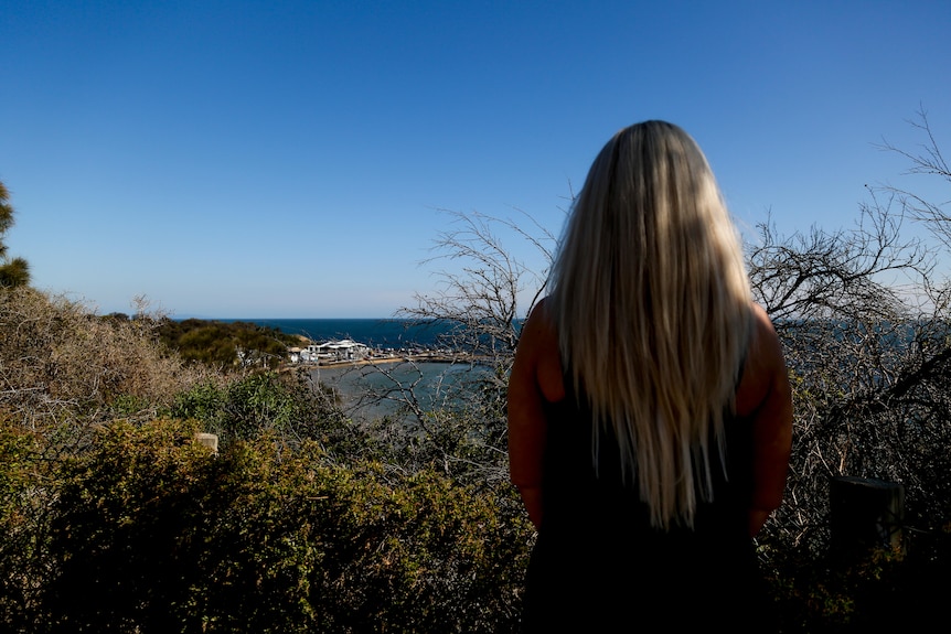 A woman with long blonde hair stands with her back to the camera, looking out towards the water. 