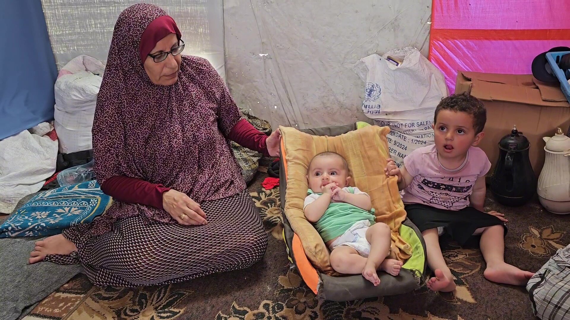 A woman wearing a purple hijab sitting on the floor with a baby and toddler next to her
