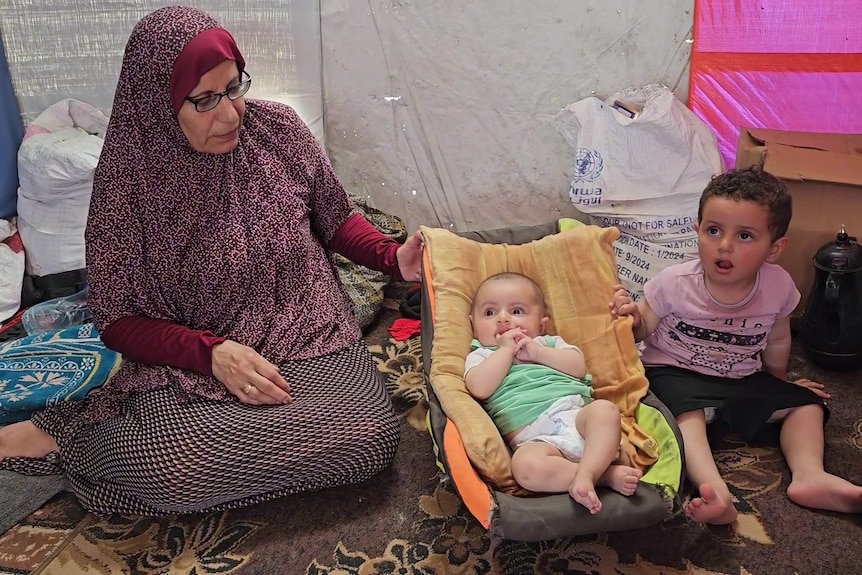 A woman wearing a purple hijab sitting on the floor with a baby and toddler next to her