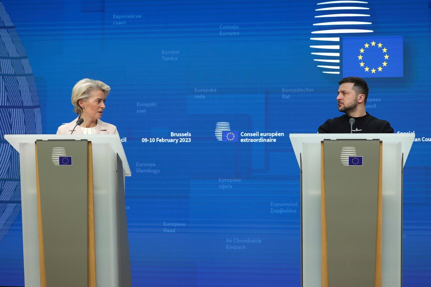 Zelenskyy and von der Leyen look at each other as they stand behind lecterns in front of a backdrop of the EU flag