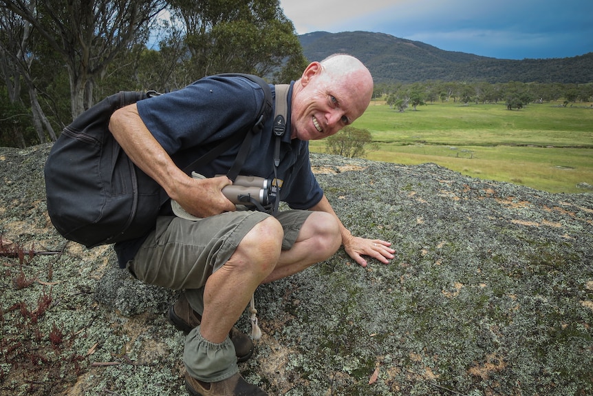 A bald man wearing shorts and tshirts places a hand on lichen covering a flat rock. 