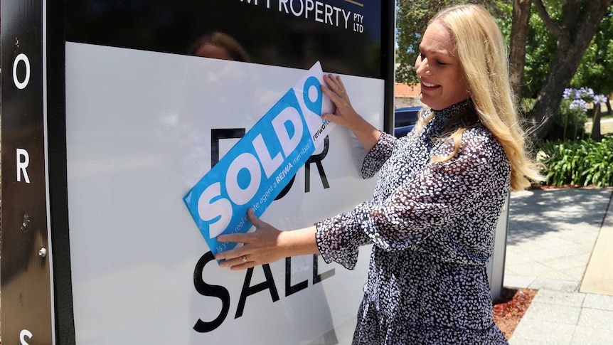 Real estate agent Chelsey Grant next to a "For Sale" sign placing a blue "SOLD" sticker over the top of it.