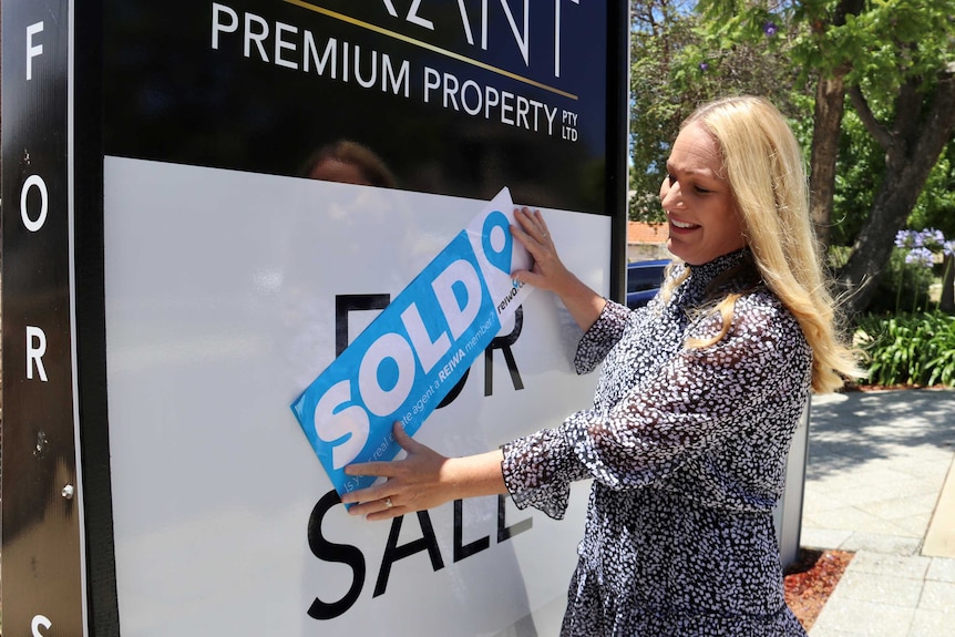 Real estate agent Chelsey Grant next to a "For Sale" sign placing a blue "SOLD" sticker over the top of it.