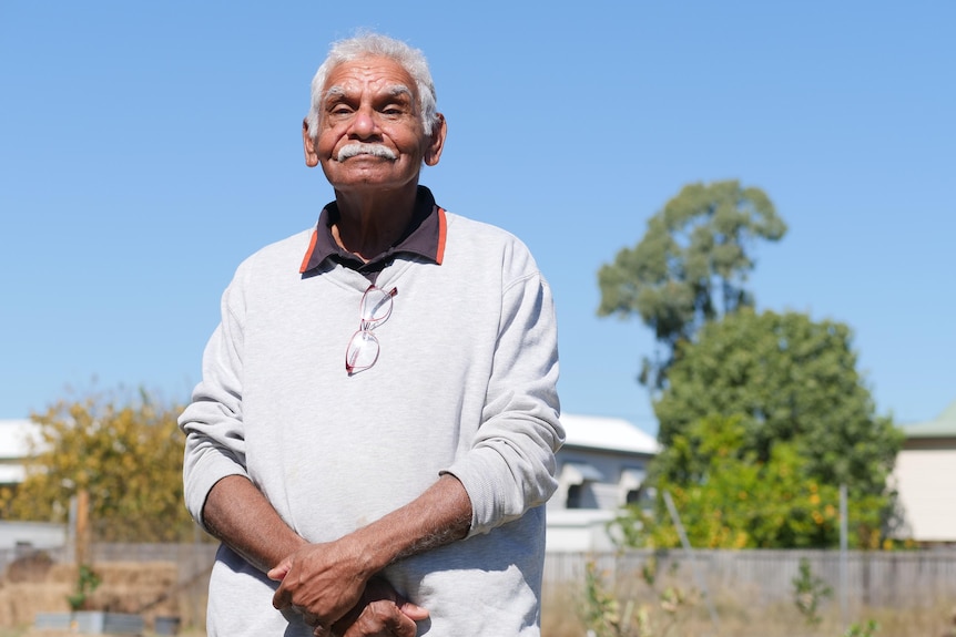 A man with grey hair and a grey moustache stands outside in the sunshine and looks at the camera. 