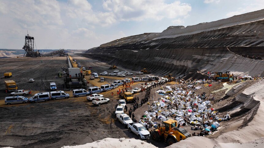 A wide view shows a large coalmine with protesters and cars lining the edges of a pit.