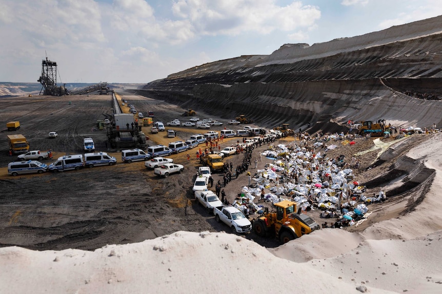 A wide view shows a large coalmine with protesters and cars lining the edges of a pit.