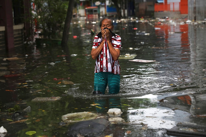 A woman holds a black scarf to her face as she wades knee-deep through rubbish-riddled floodwater.
