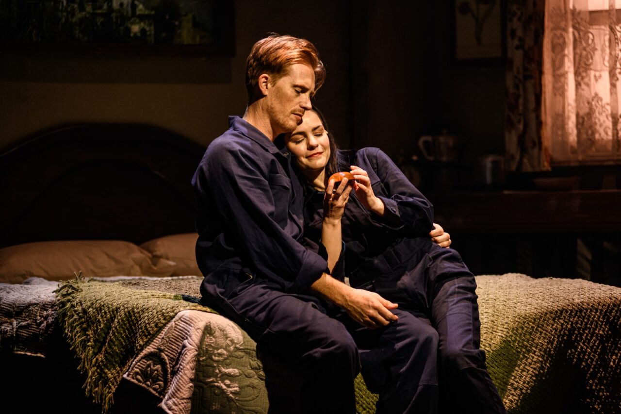 A male and female actor embrace sitting on a bed.