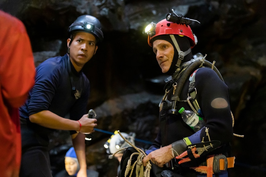一名身穿海军衬衫和头盔的泰国男子手持火炬，旁边是一名身穿潜水装备和头灯的白人男子，在黑暗的洞穴中