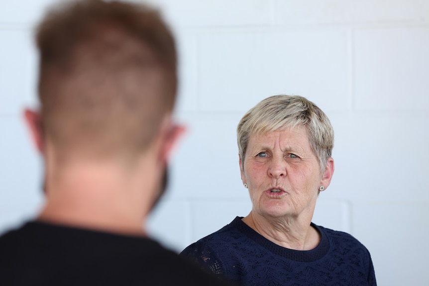 A woman with short blonde hair speaks to a man in the foreground.