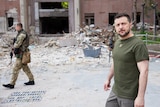 An armed soldier and a man walk along a destroyed city street filled with rubble.
