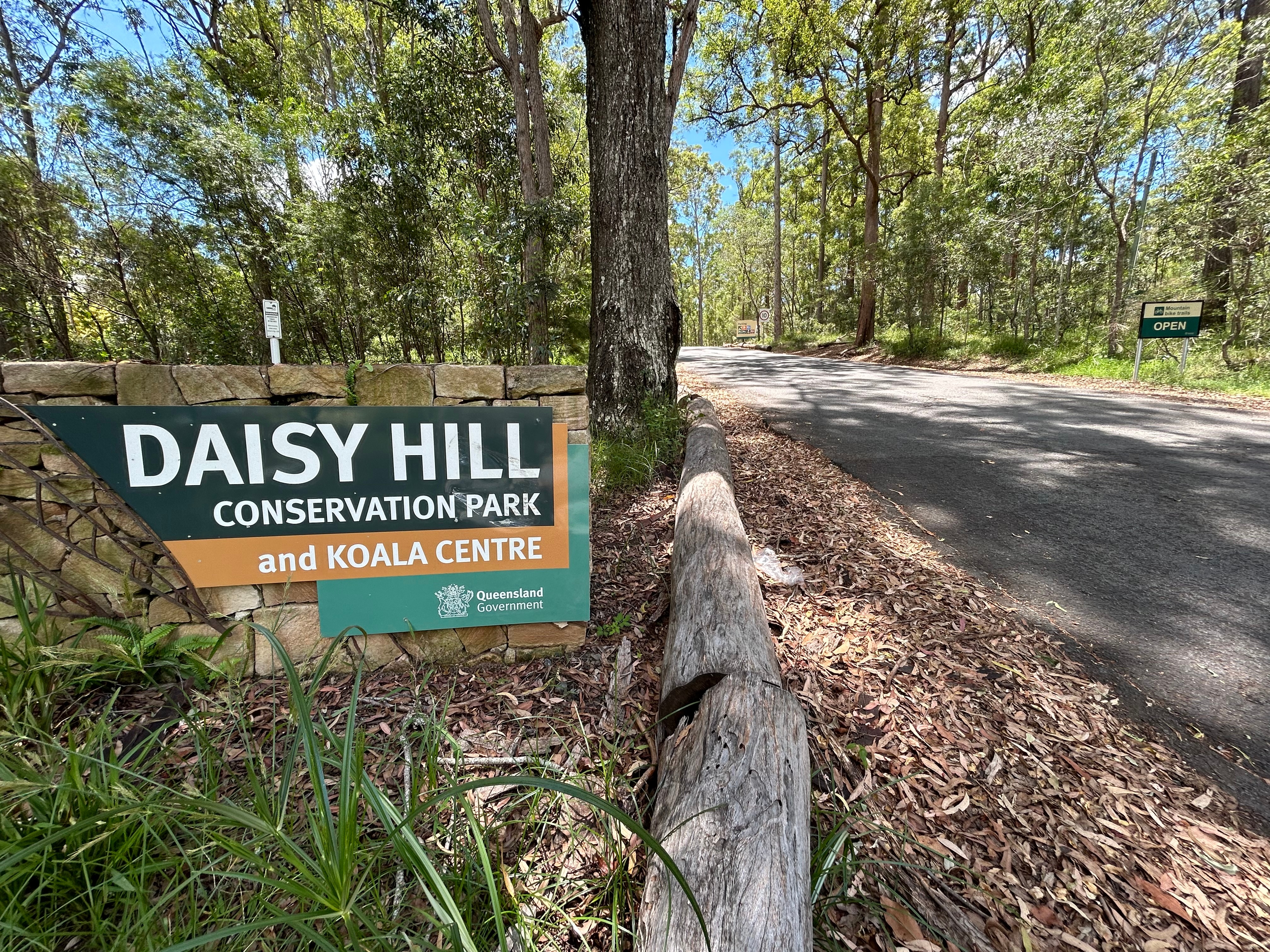 A picture of a sign that says 'Daisy Hill Conservation Park and Koala Centre' with trees and a road in the background.