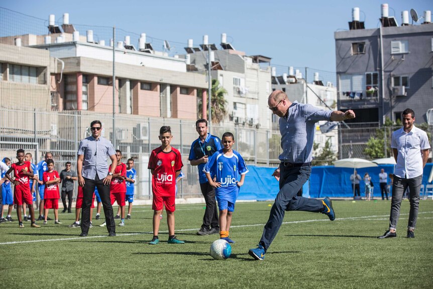 Prince William plays soccer with Jewish and Arab children. He is kicking the ball.