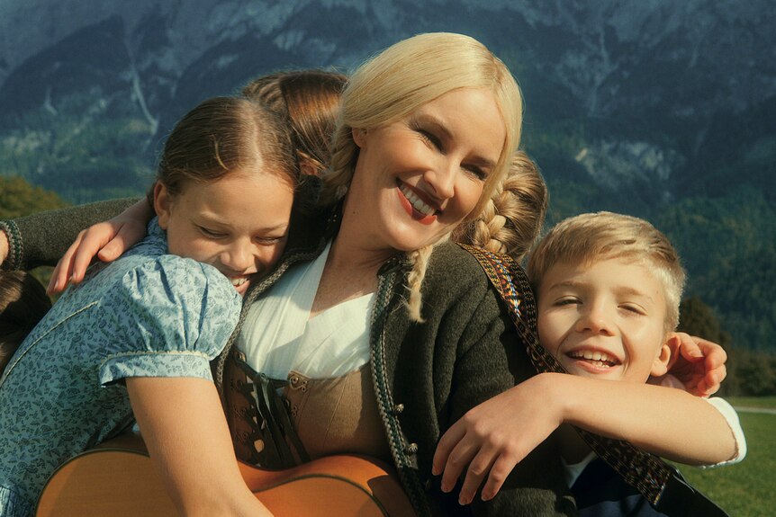 A blonde woman in her 40s, her hair in plaits, smiles brightly while hugging a group of blonde children in Austrian countryside