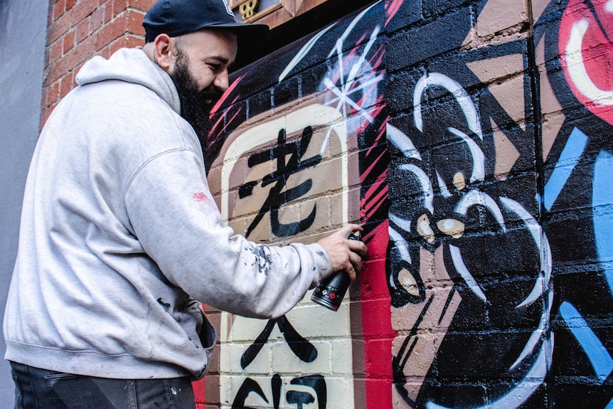 A man in a hat and jumper sprays paint onto a brick wall.