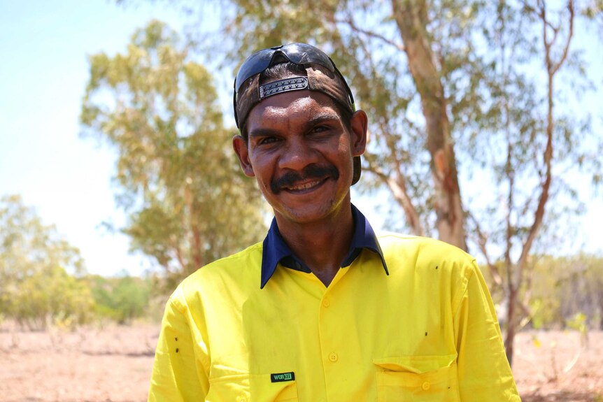 An indigenous man in high visibility work shirt, wearing a backwards cap in front of bushland