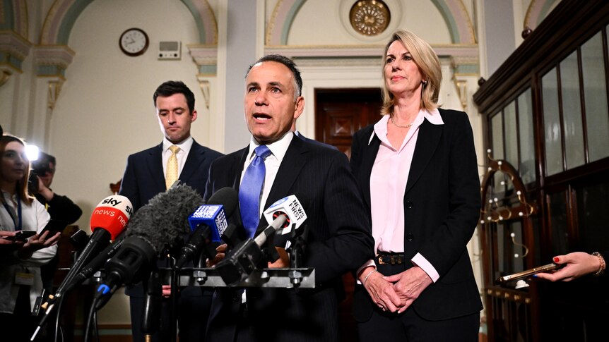 A man in a suit, centre, flanked by two other people, standing in front of microphones and members of the press.