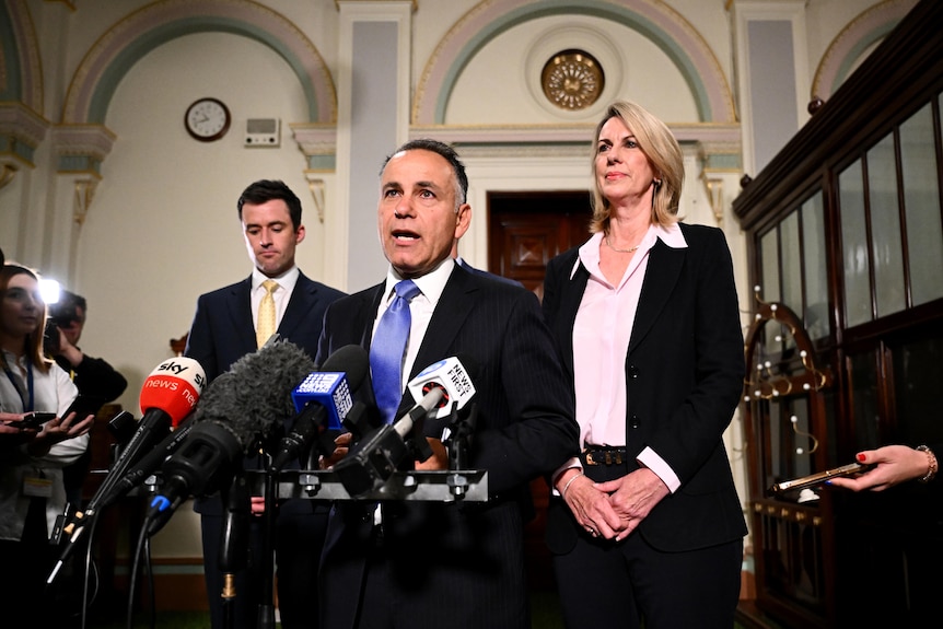 A man in a suit, centre, flanked by two other people, standing in front of microphones and members of the press.