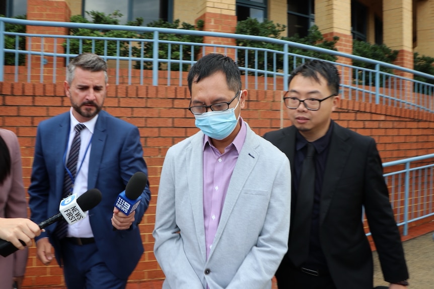 Tsz Wing Tam in a grey suit jacket and a face mask, leaving court surrounded by journalists.