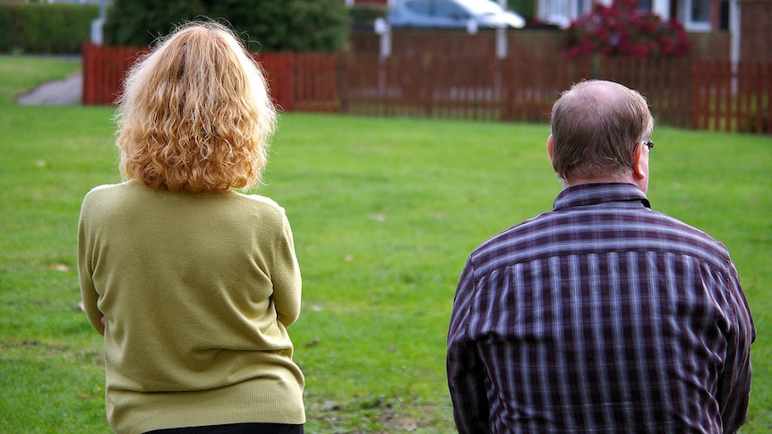 A young woman and older man stand beside each other in front of a picketed fence house