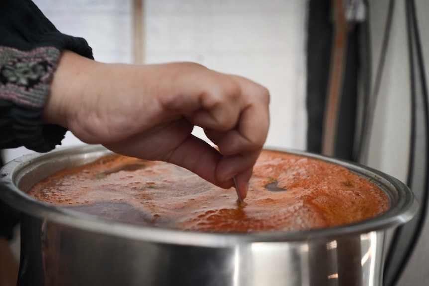 A woman's hand stirs a red-orange broth in a stainless steel pot