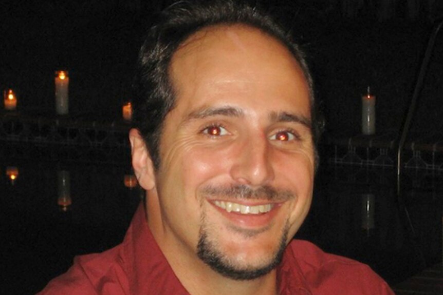 A balding man with a thin goatee smiles as he looks into the camera while standing before a row of candles.