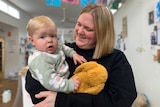 A woman wearing a black jumper holds a child wearing a green jumper.