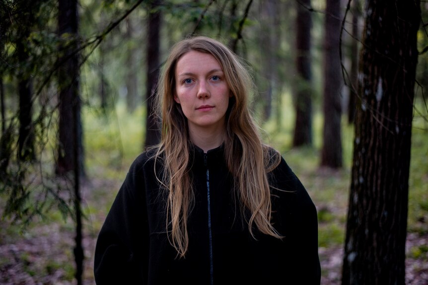 A woman with long hair wears and hoodie and stands in the forest. 