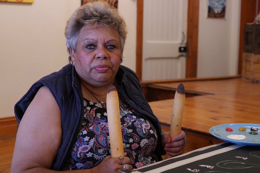 An Aboriginal woman with short grey hair sits at a table in front of her painting holding traditional clapsticks.