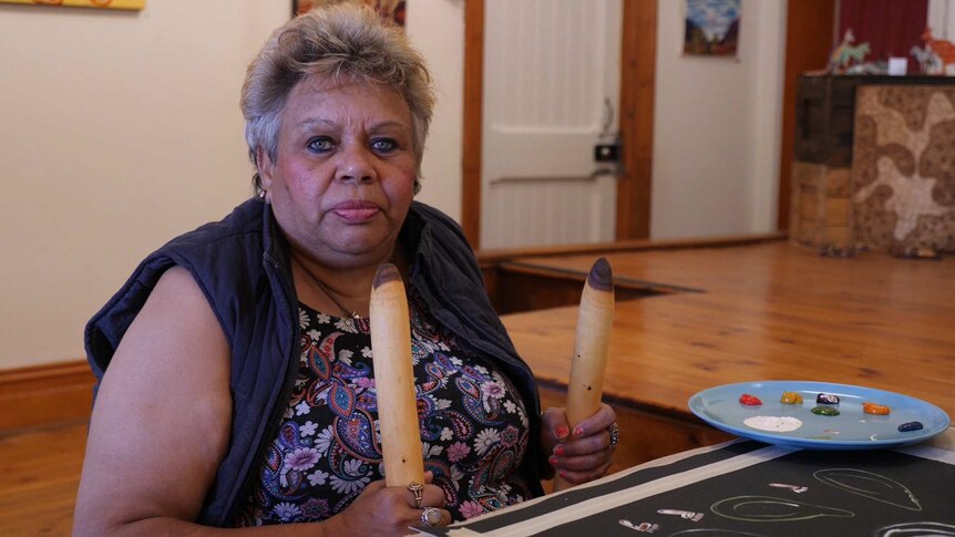 An Aboriginal woman with short grey hair sits at a table in front of her painting holding traditional clapsticks.