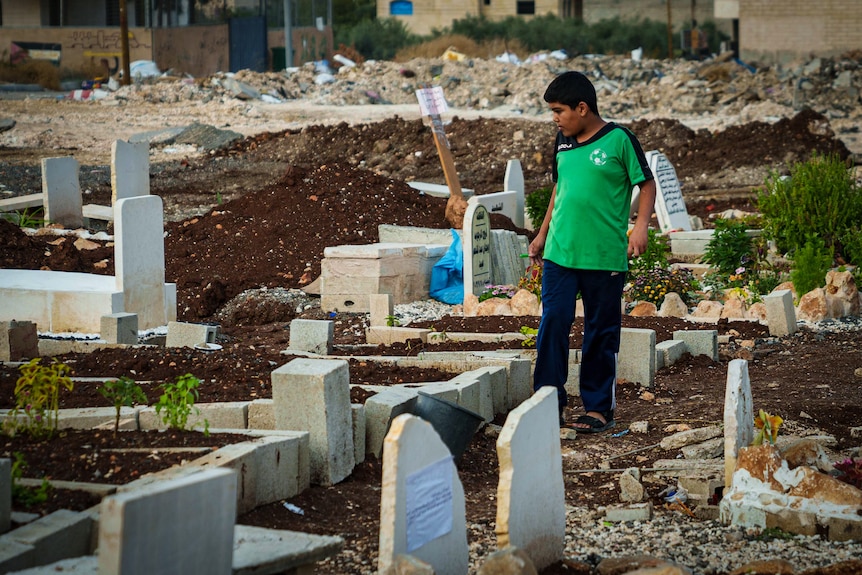 A boy walks along a line of graves in a graveyard.